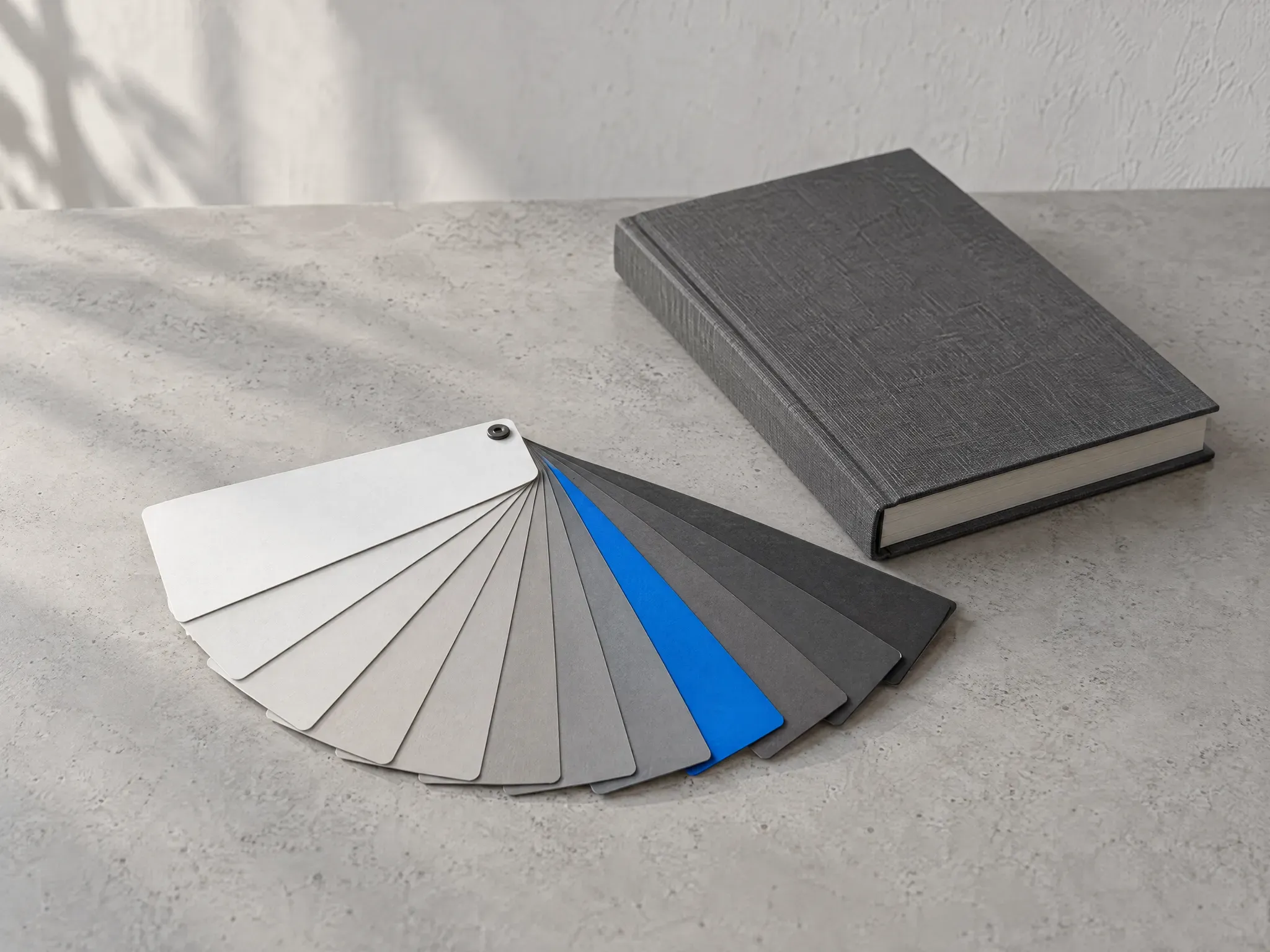 A fan of grey paper colour swatches with one blue swatch, beside a closed charcoal hardback book on pale concrete